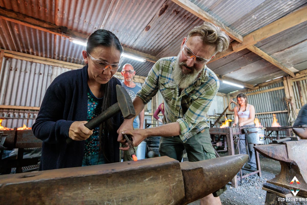 Blacksmith instructor guiding a student with hammer and tongs